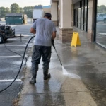 Worker pressure washing a shopping center sidewalk next to a trailer-mounted pressure washer