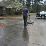 Worker pushing a surface cleaner across a concrete driveway in front of a brick ranch home with a flowering dogwood